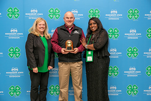 A woman, a man, and another woman stand in front of a blue background with the 4-H and MSU Extension logos repeated across