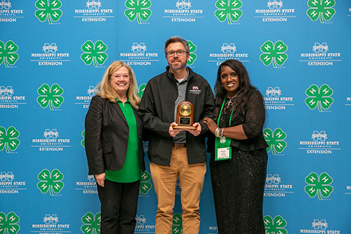 A woman, a man, and another woman stand in front of a blue background with the 4-H and MSU Extension logos repeated across