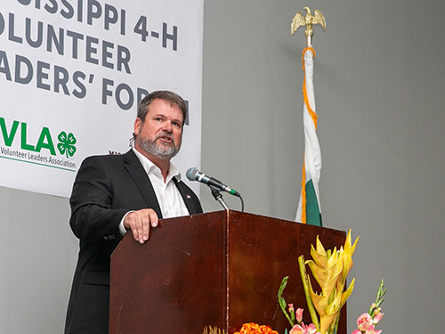 A man stands at a podium speaking in front of a flag and MVLA banner