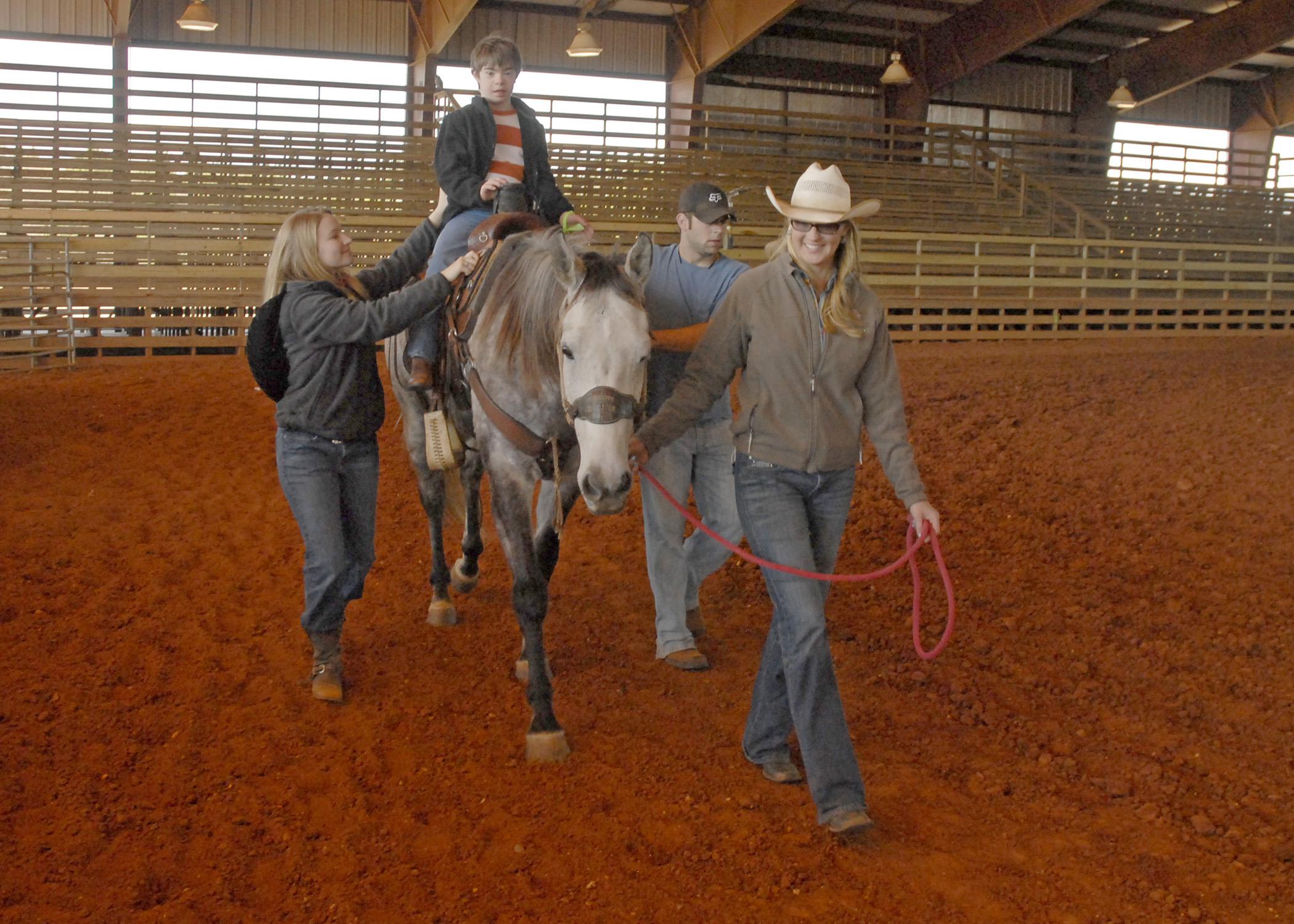 A child rides a horse as two adults prevent him from falling and a man leads the horse.